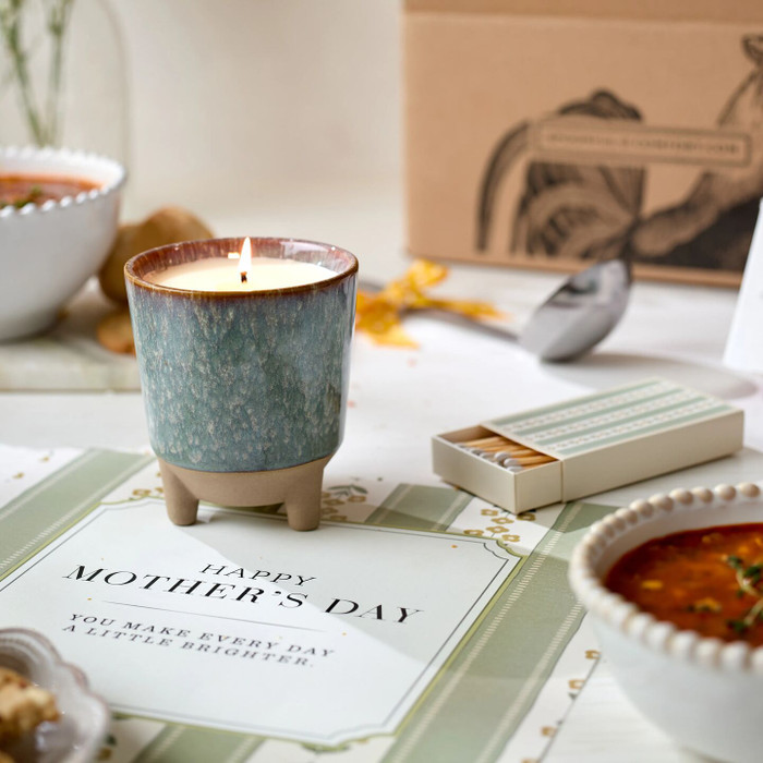 Close-up of a lit candle on a Mother’s Day card, with a matchbox, a bowl of soup, and a ladle in the background.