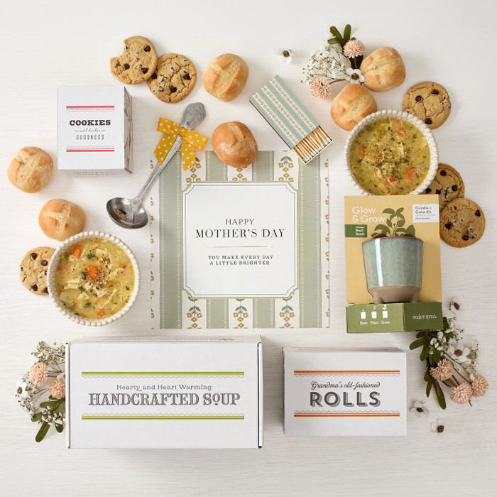 Overhead view of a Mother’s Day gift arrangement with two bowls of soup, rolls, cookies, a box labeled “Handcrafted Soup,” a box of rolls, a candle grow kit, a metal ladle with a yellow ribbon, and a card that reads “Happy Mother’s Day.”