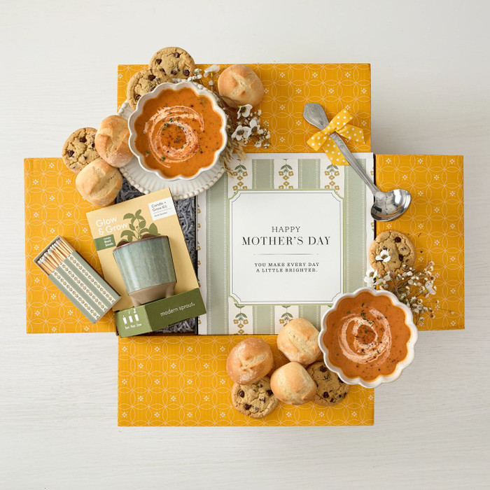 Overhead view of a yellow gift box arrangement with two bowls of soup, rolls, cookies, a candle gift set, a metal ladle with a yellow ribbon, and a card that reads “Happy Mother’s Day.”