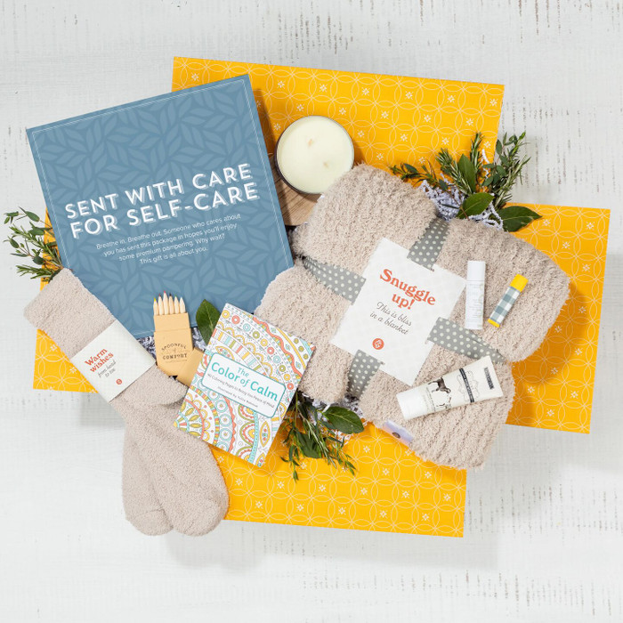 Overhead view of a yellow gift box containing a blue card that reads “Sent With Care for Self-Care,” a candle, one pair of socks, small self-care items, and greenery.