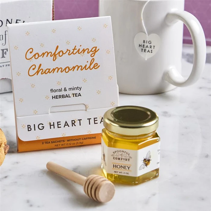 Close-up of a package labeled “Comforting Chamomile” herbal tea next to a mini jar of honey with a wooden dipper, with a mug in the background.