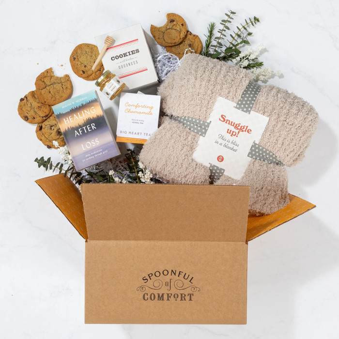 Open Spoonful of Comfort gift box filled with a cozy knit blanket labeled “Snuggle Up,” cookies, a box of cookies, a package of chamomile tea, a jar of honey with a dipper, and a book titled “Healing After Loss,” arranged with greenery.