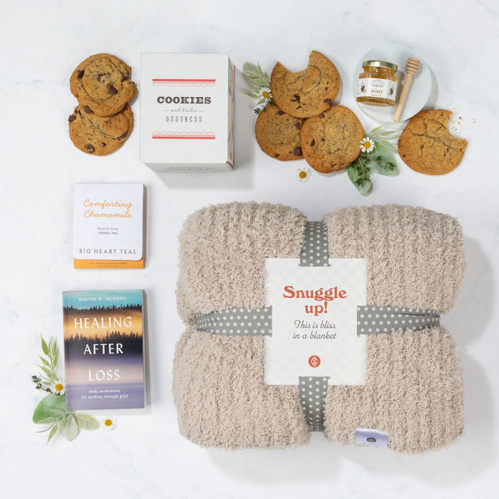 Overhead view of a yellow gift box containing a cozy knit throw blanket, a box of cookies, assorted cookies, tea sachets, a jar of honey with a honey dipper, a healing-after-loss book, and greenery.