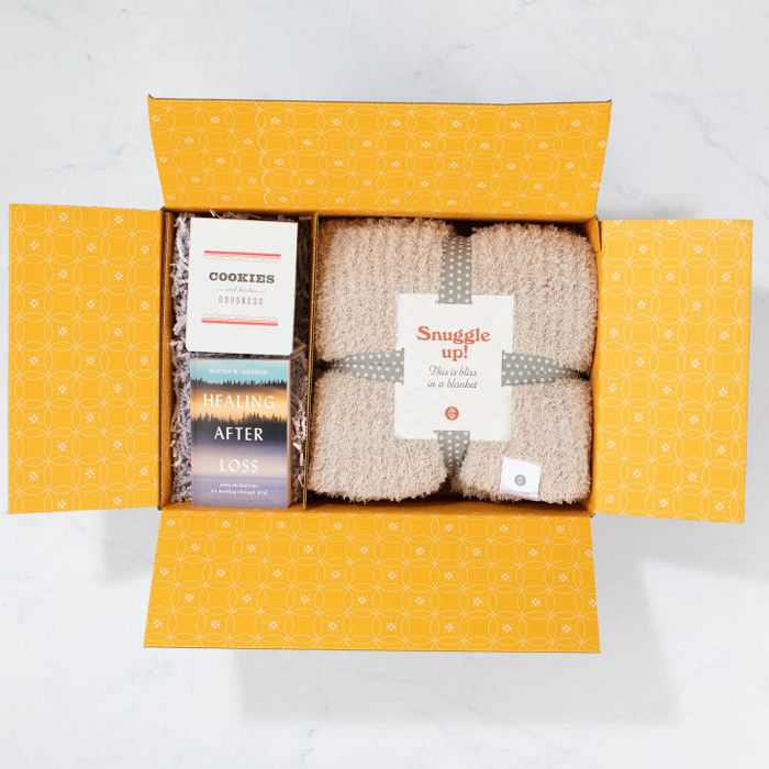 Open yellow gift box displaying a cozy knit blanket labeled “Snuggle Up,” alongside a box of cookies and a book titled “Healing After Loss.”