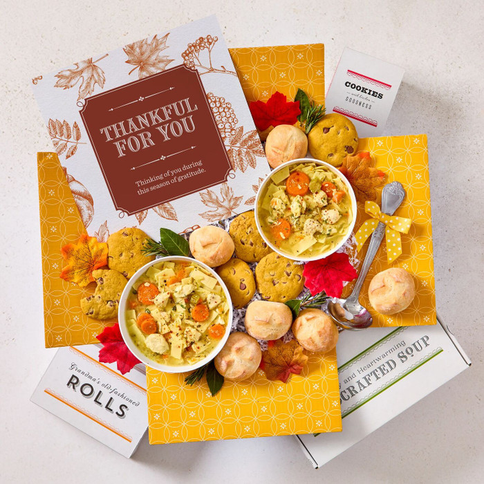 Overhead view of a fall-themed gift arrangement with two bowls of soup, rolls, cookies, a container labeled “Handcrafted Soup,” a metal ladle with a yellow ribbon, autumn leaves, and a card that reads “Thankful for You.”
