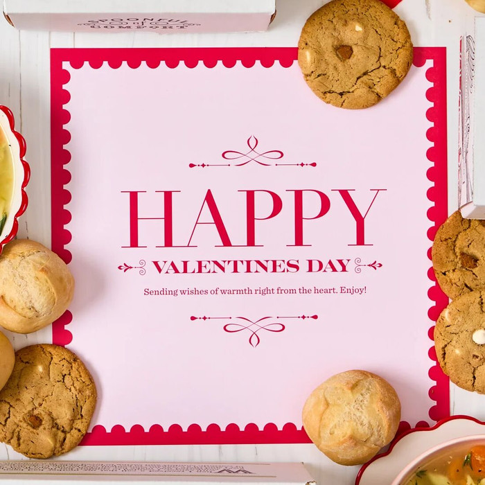 Overhead view of hands holding a Valentine’s-themed gift arrangement with two bowls of soup, rolls, cookies, a container labeled “Handcrafted Soup,” heart-shaped decorations, and cards that read “Happy Valentine’s Day”
