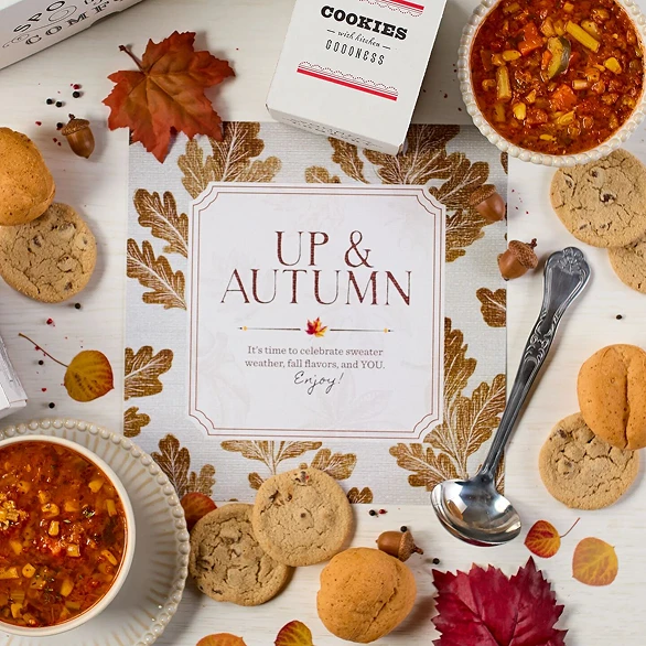 Overhead view of hands holding a fall-themed gift arrangement with two bowls of soup, rolls, cookies, a container labeled “Handcrafted Soup,” a metal ladle with a yellow ribbon, autumn leaves, and a card that reads “Up and Autumn.”