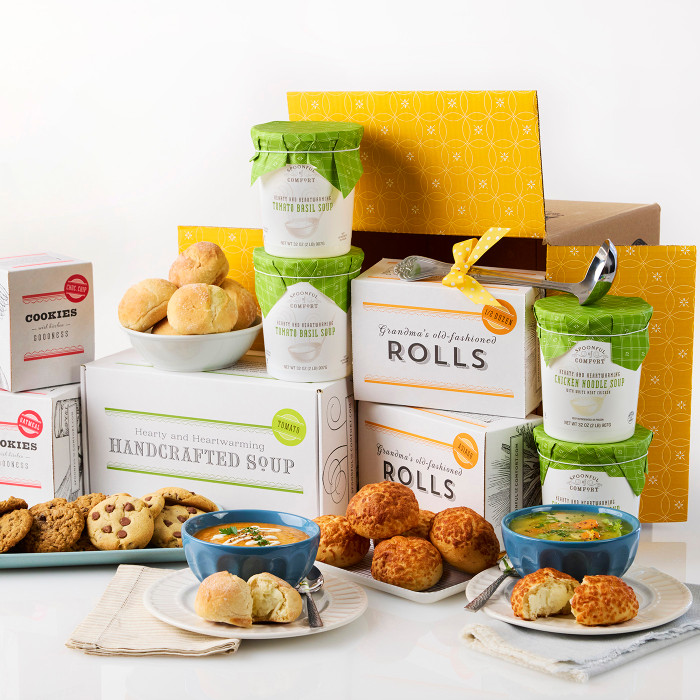 Soup gift set displayed on a table with bowls of soup, rolls, cookies, multiple containers of soup with green lids, boxes labeled “Handcrafted Soup” and “Rolls,” and an open yellow gift box in the background.