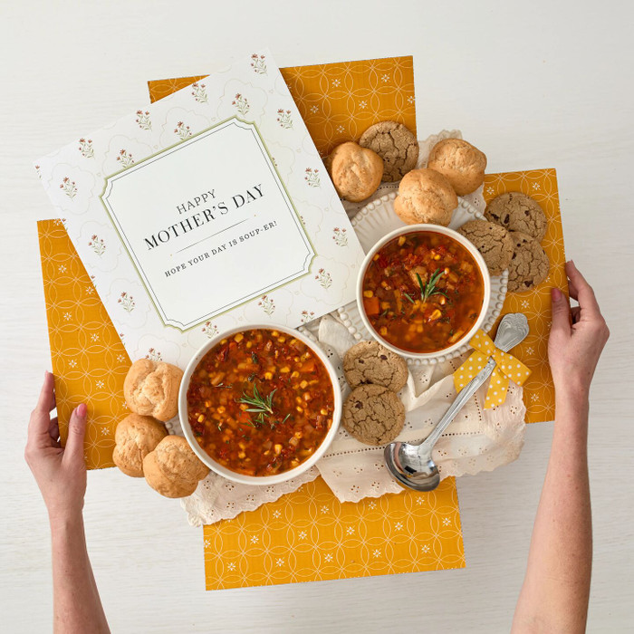 Overhead view of hands holding a yellow gift box arrangement with two bowls of soup, rolls, cookies, a metal ladle with a yellow ribbon, and a card that reads “Happy Mother’s Day.”