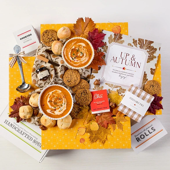 Overhead view of a fall-themed gift arrangement with two bowls of soup, rolls, a container labeled “Handcrafted Soup,” a metal ladle with a yellow ribbon, autumn leaves, and a card that reads “Up and Autumn.”