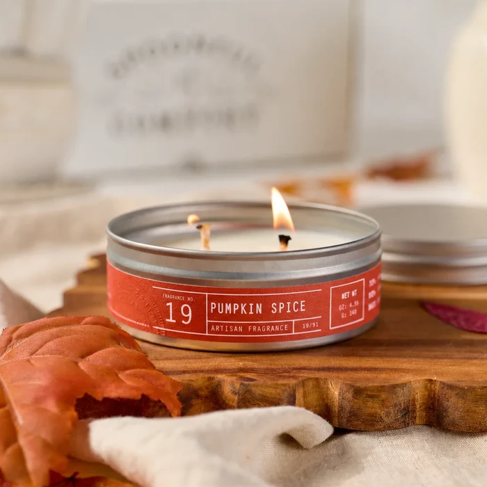 Lit pumpkin spice candle in a red tin on a wooden cutting board, with autumn leaves and a folded cloth nearby.