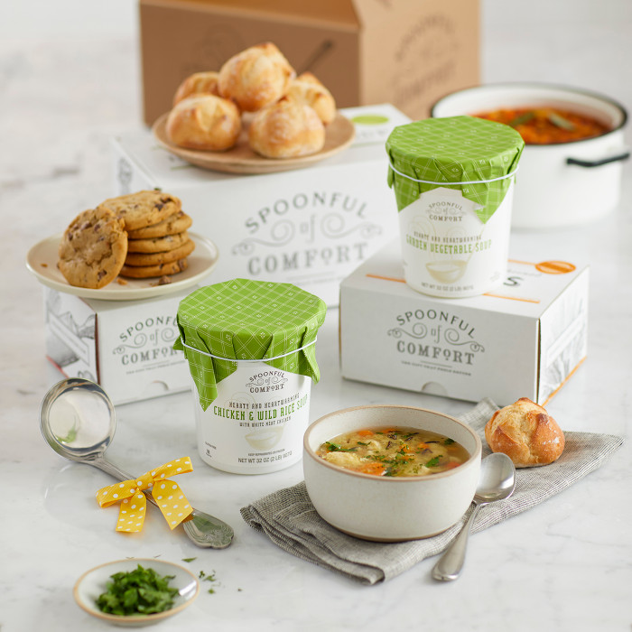Soup gift set on a counter with bowls of soup, rolls, cookies, two containers of soup with green lids, a metal ladle with a yellow ribbon, and Spoonful of Comfort boxes in the background.
