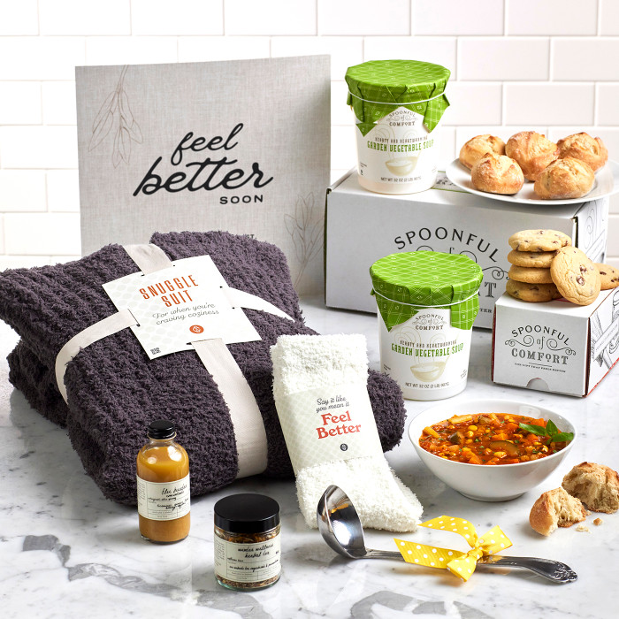 Comfort-themed gift set on a counter with a knit blanket, fuzzy socks, a bowl of soup, rolls, cookies, two containers of soup, a candle, small self-care items, a metal ladle with a yellow ribbon, and a sign that reads “Feel Better.”