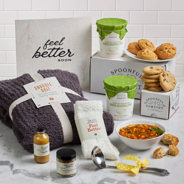 Comfort-themed gift set on a counter with a knit blanket, fuzzy socks, a bowl of soup, rolls, cookies, two containers of soup, a candle, small self-care items, a metal ladle with a yellow ribbon, and a sign that reads “Feel Better.”