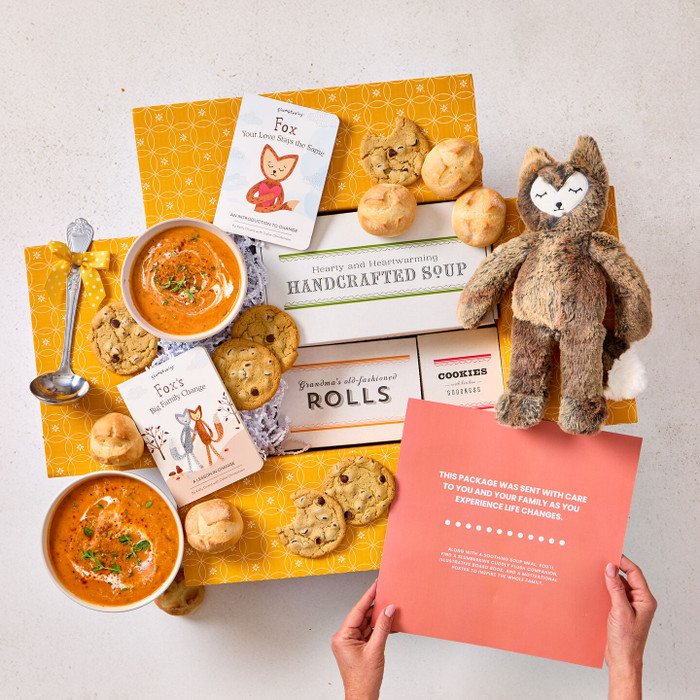 Overhead view of hands holding a card with a heartfelt message next to a comforting gift arrangement with two bowls of soup, rolls, cookies, a box labeled “Handcrafted Soup,” a box of rolls, a plush stuffed animal, children’s books, and a metal ladle with a yellow ribbon.