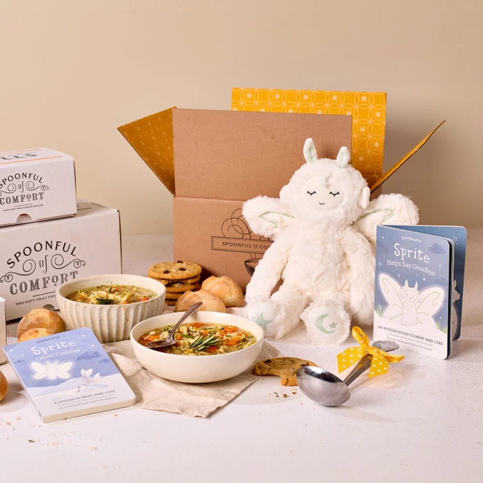 Comfort-themed gift set displayed with bowls of soup, rolls, cookies, Spoonful of Comfort boxes, a plush Sprite toy, children’s books, and a metal ladle with a yellow ribbon.
