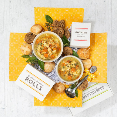 Overhead view of a yellow gift box arrangement with two bowls of soup, rolls, cookies, a box of cookies, a container labeled “Handcrafted Soup,” and a metal ladle.