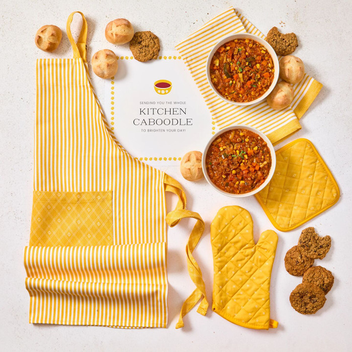 Overhead view of a kitchen-themed arrangement with two bowls of soup, rolls, cookies, a striped apron, matching oven mitts, and a card that reads “Kitchen Caboodle.”