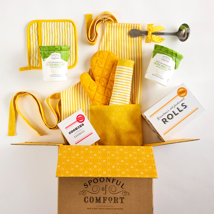 Open Spoonful of Comfort gift box filled with yellow kitchen items including a striped apron, oven mitts, towel, utensils, packaged soup containers, cookies, and a box of rolls.
