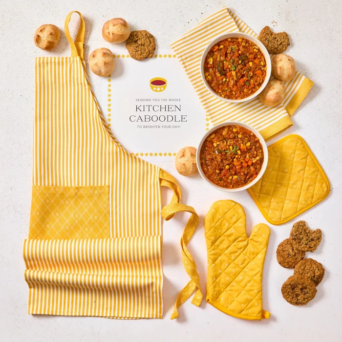 Overhead view of a kitchen-themed arrangement with two bowls of soup, rolls, cookies, a yellow striped apron, matching oven mitts and pot holder, and a card that reads “Kitchen Caboodle.”