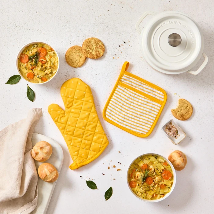 Overhead view of a kitchen-themed arrangement with two bowls of soup, rolls, cookies, a yellow oven mitt and pot holder, a white Dutch oven lid, and a towel on a light surface.