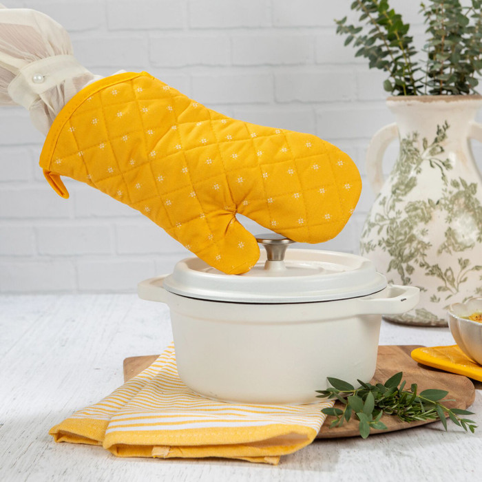 Hand wearing a yellow oven mitt lifting the lid of a white Dutch oven on a wooden board, with a striped towel and greenery nearby.
