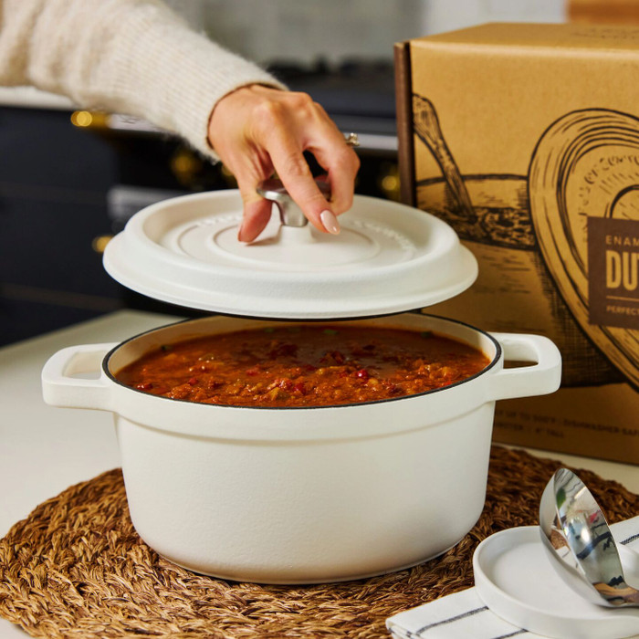 Hand lifting the lid of a white Dutch oven to reveal soup inside, with the product box and a spoon nearby on a table.