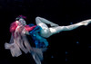 Underwater photograph by Caroline Gray depicting a woman floating gracefully in a dark pool, wearing sheer pink fabric and vibrant blue layers, with a floral crown adorning her head and dramatic lighting illuminating her pale form.