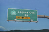 Photorealistic oil painting of a Laguna Canyon Road exit sign floating against a bright blue sky above distant dark green hills. Laguna Canyon by Eric Nash, Oil on canvas, 48” x 72” 2026, $16,500