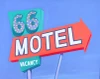 Joan Gladstone’s painting depicts a vintage Route 66 motel sign in Needles, California, featuring an illuminated “66,” a bold red-orange arrow with white MOTEL lettering, and a turquoise “No Vacancy” sign against a luminous blue sky.