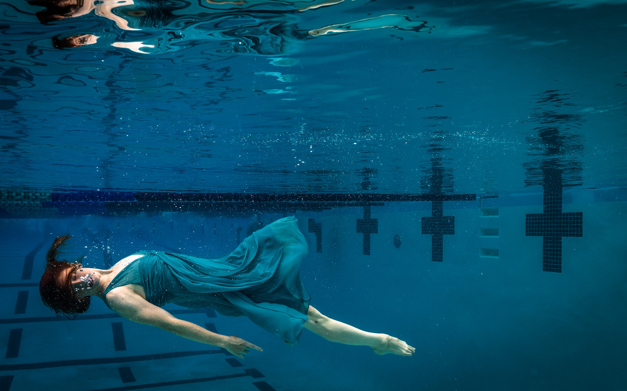 Underwater photograph by Caroline Gray featuring a woman in a flowing teal dress gliding horizontally through a pool, with bubbles rising from her mouth and soft light illuminating her figure against the geometric lines of the pool floor and walls.