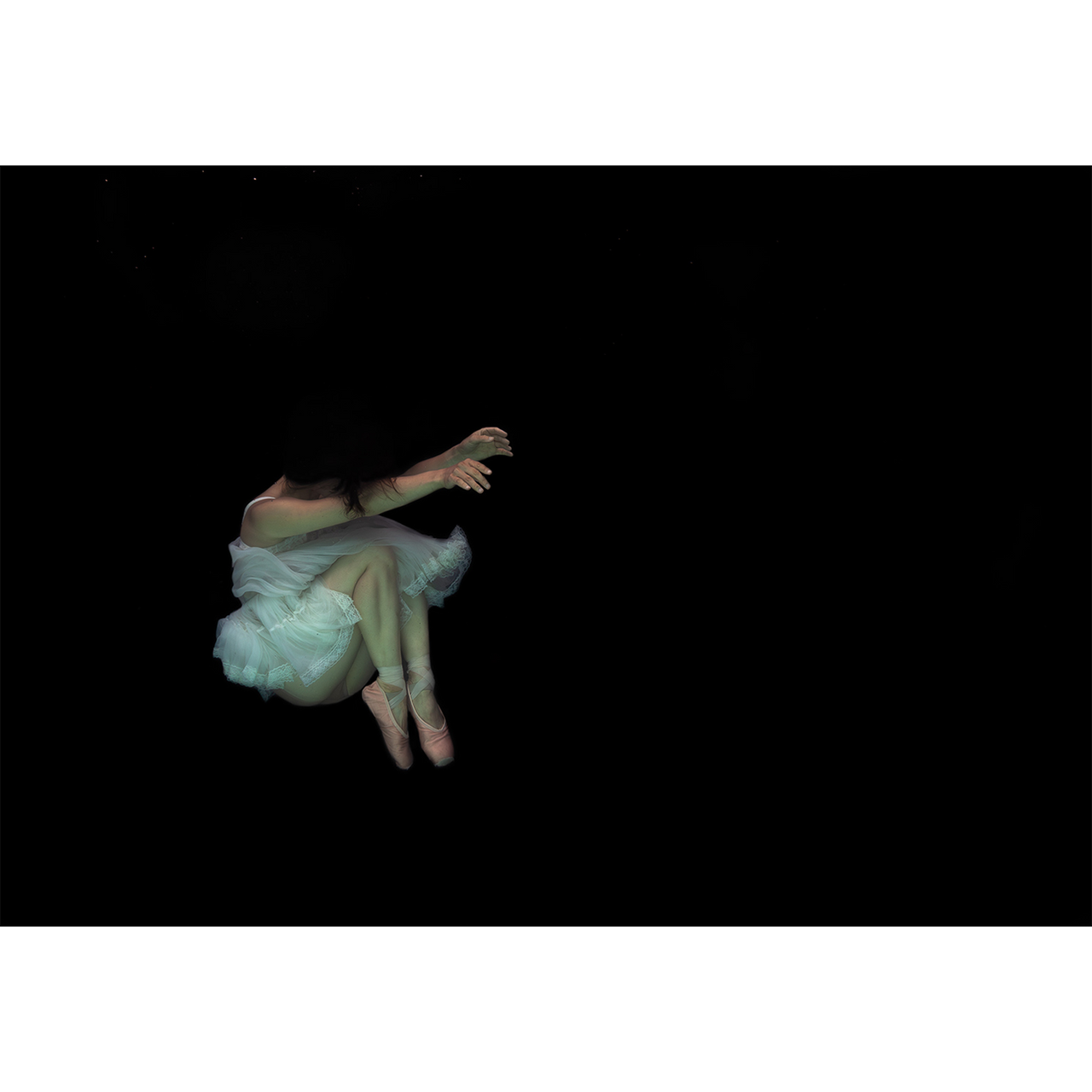 Underwater photograph by Caroline Gray depicting a ballerina in a white tutu and pale pink pointe shoes suspended in a vast black expanse of water, her body curled inward with arms gently extended, evoking quiet introspection and weightless stillness.