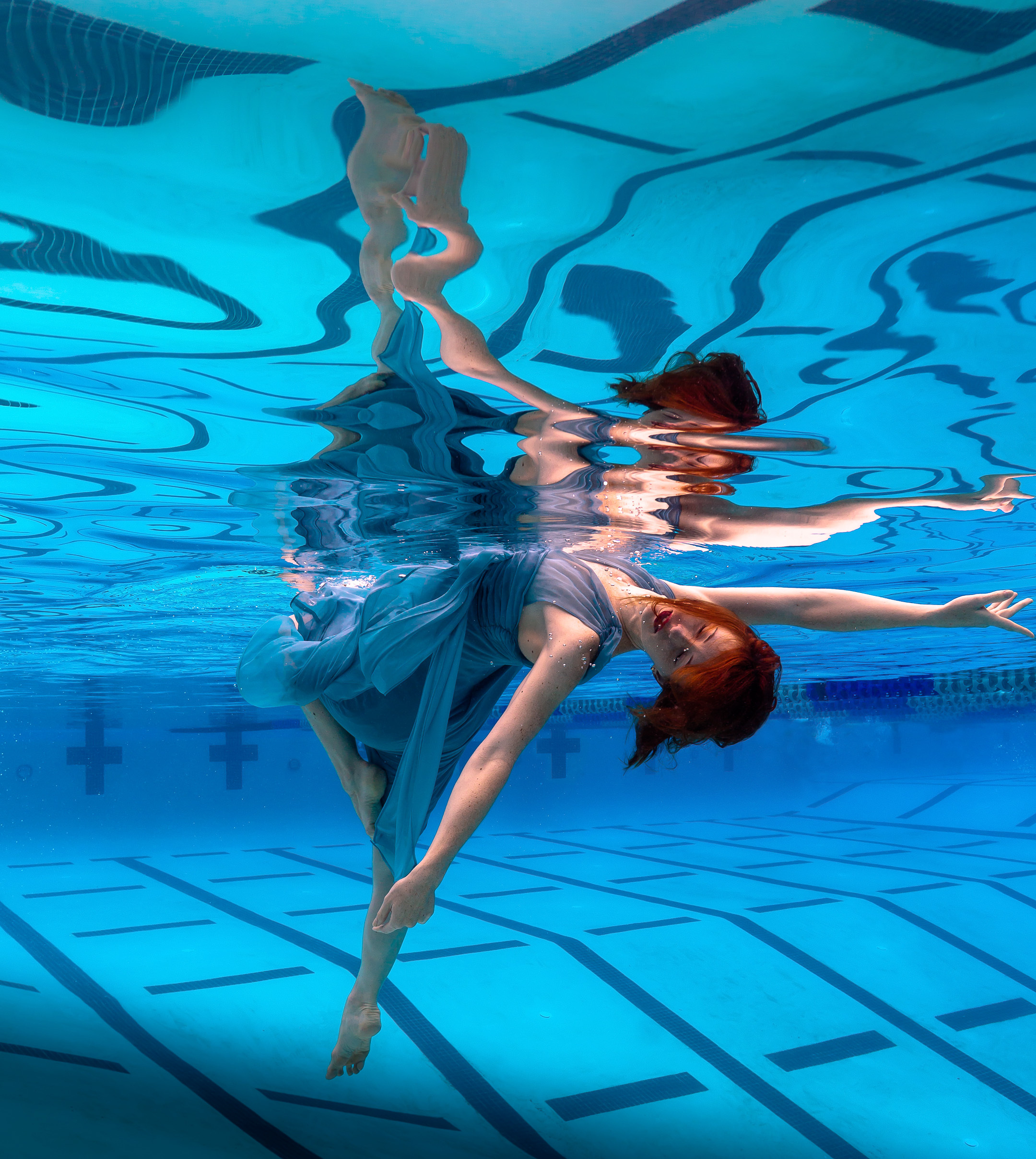 Underwater photograph by Caroline Gray featuring a woman in a flowing blue dress gracefully suspended below the water&rsquo;s surface, with her reflection rippling above in a serene pool setting.