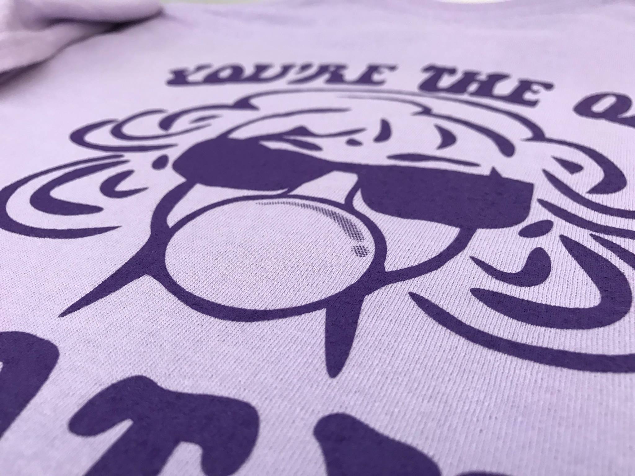 A light purple t-shirt with a dark purple screen print of a woman blowing a bubble with bubble gum while wearing sunglasses.