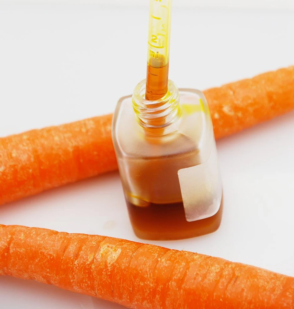 A dropper bottle of golden carrot-seed oil resting on fresh carrots on white, evoking natural vitamin A (beta-carotene) for skin