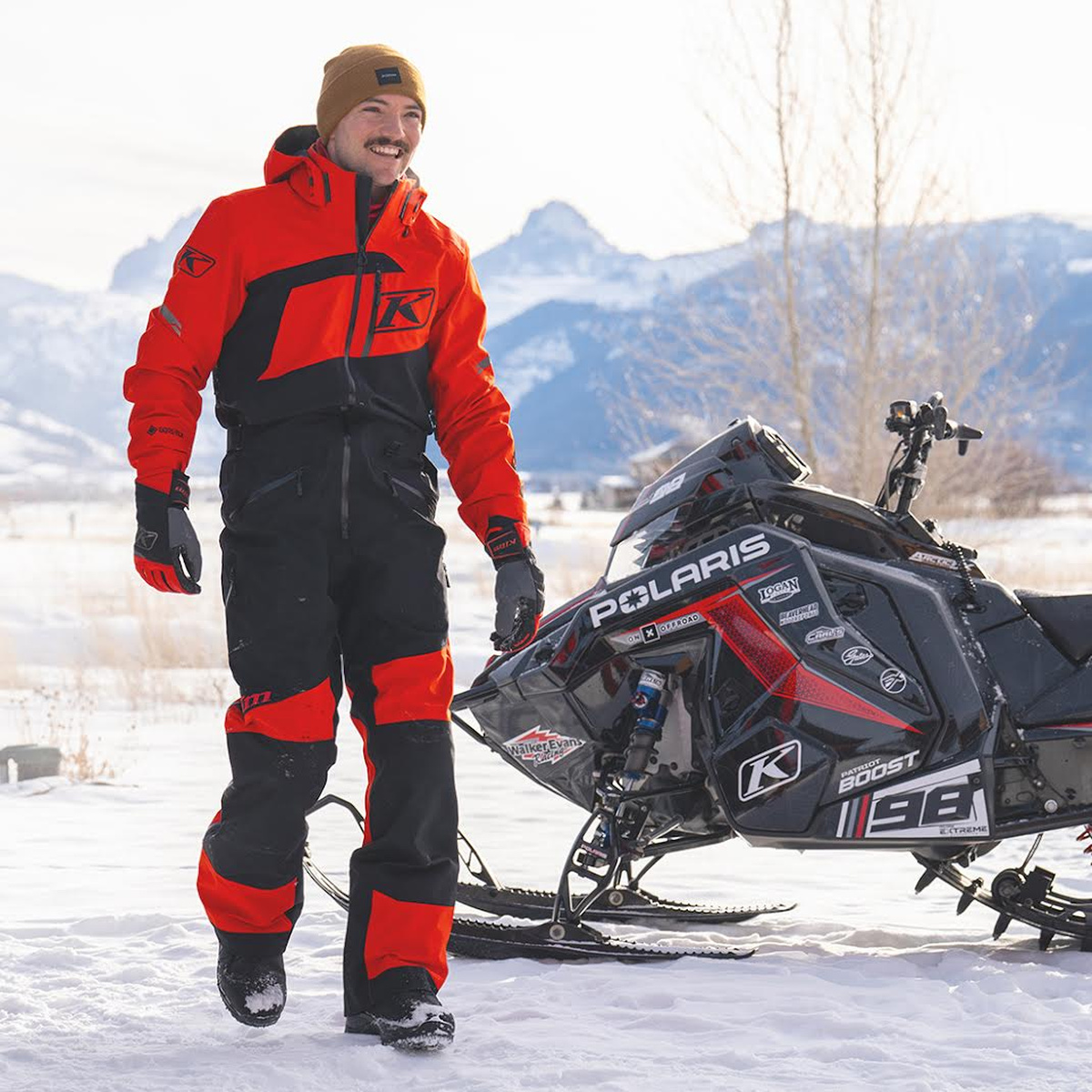 Rider wearing a red Klim snow suit beside a snowmobile
