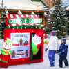 Close-up of Santa Claus standing in front of red gingerbread cookie shop inflatable, smiling and holding a candy cane in festive holiday display
