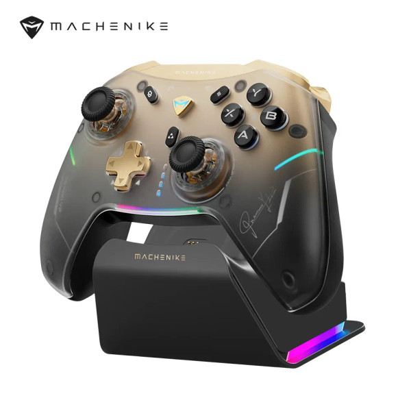 Gold MACHENIKE G5Pro V2 gaming controller glowing with customizable RGB lighting on a dark desk setup with keyboard and monitor in the background.