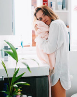 A young woman in a white shirt holds a pink towel in a bright bathroom setting with plants and toiletries.