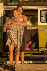 A man and a woman embrace, wrapped in a black and white patterned beach towel, beside a vintage van. A man and a woman embrace, wrapped in a black and white patterned beach towel, beside a vintage van.