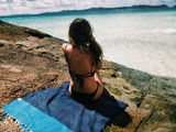 A beach towel in navy blue with a patterned edge laid on a rock, with a person sitting on it, facing the water. A beach towel in navy blue with a patterned edge laid on a rock, with a person sitting on it, facing the water.