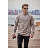 A man wearing a beige crew neck fleece sweater and sunglasses, holding a camera while walking on a pier.