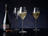 A drink bottle alongside three elegant clear glass Prosecco glasses filled with sparkling beverage, set against a dark background.