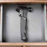 A black survival hammer with multiple tools, displayed in a grey drawer. It features a sleek design and a branding logo.