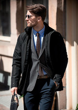A stylish black men's jacket worn over a grey blazer with a light blue shirt, paired with sunglasses and trousers.