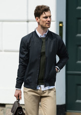 A man wearing a black bomber jacket over a green sweater, carrying a brown bag, standing near a green door.