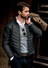 A man wearing a black quilted jacket, holding a brown bag, with sunglasses and a casual shirt underneath.