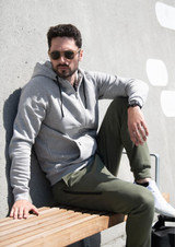 A grey hoodie jacket sits on a man lounging on a wooden bench, wearing green pants and sunglasses.