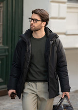 A man in a black jacket walks outside, wearing glasses and carrying a bag, with a green top and light trousers.