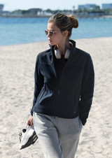 A women’s navy jacket with a high collar, paired with grey pants, worn by a model on a sandy beach.